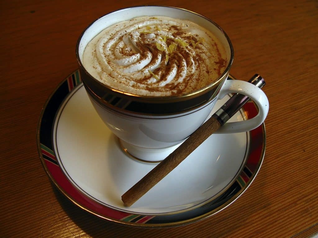 Imagem de uma xícara com capuccino de canela. Ela está sobre um pires e ao lado um pedaço de canela em pau.