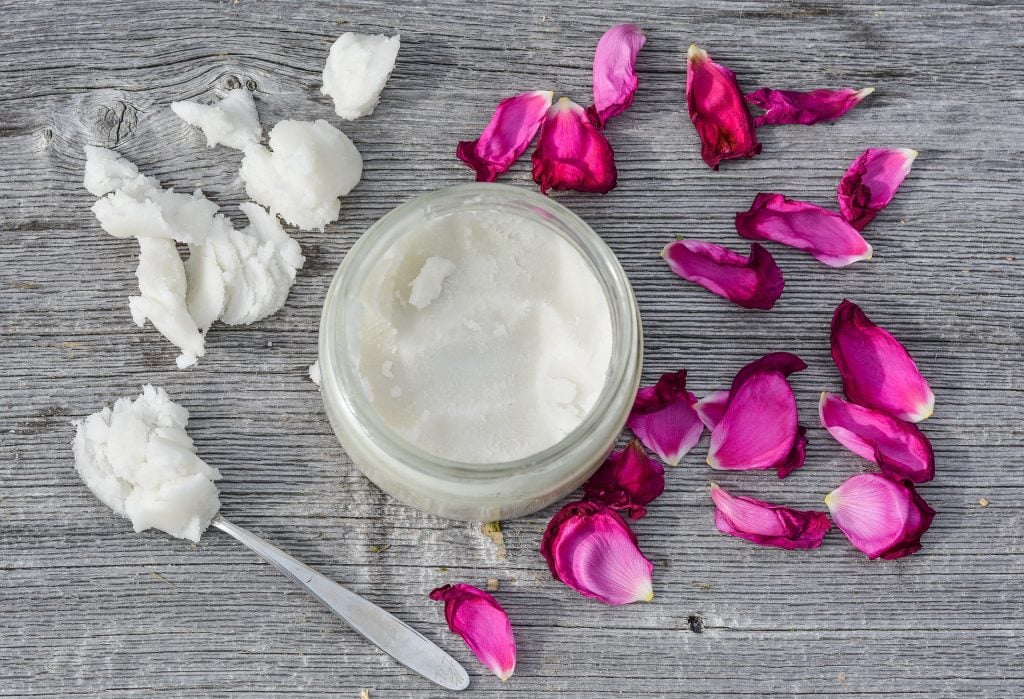 Creme para a hidratao feito  base de leo de coco. O creme est em um pote de vidro disposto sobre uma mesa de madeira cinza. Ao lado do pote ptalas de rosas rosa.