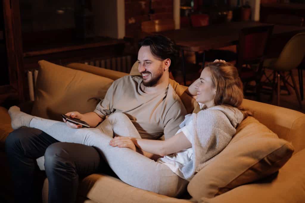 Homem e mulher sentados no sofá, assistindo à televisão enquanto riem.