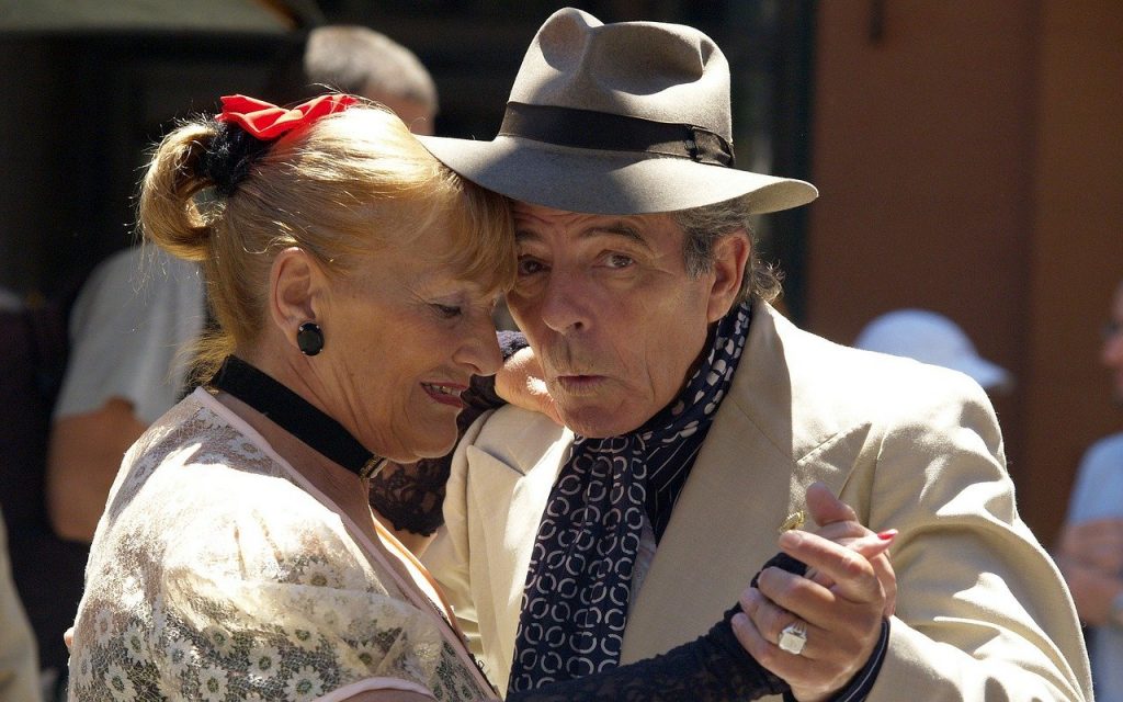 Imagem de um casal (homem e mullher) dançando tango. Eles estão felizes. O homem usa um chapéu cinza e a mulher está com o cabelo preso com um laço vermelho.