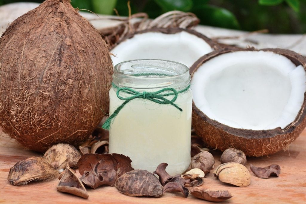 Imagem do óleo de coco em um vidro médio. Ao lado dele um coco inteiro e outro cortado ao meio e algumas castanhas espalhadas.