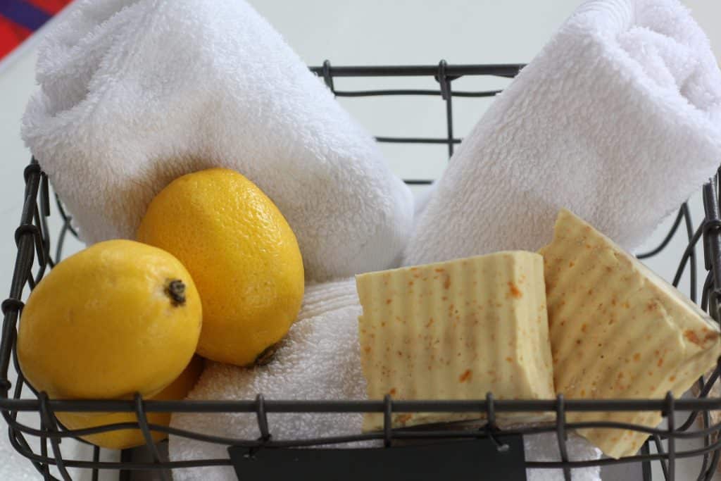 Imagem de uma cesta de arame com pedras de sabo feito de leo de coco, limes  e trs toalhas brancas de banho.