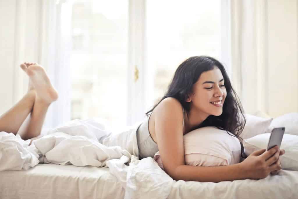 Mulher jovem deitada na cama, rindo enquanto usa o celular.