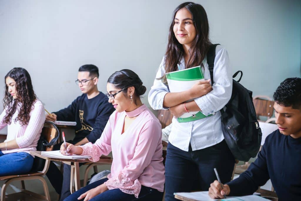 Aluna em pé na sala de aula segurando seus livros e sorrindo