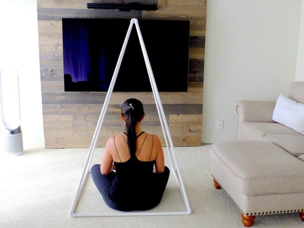 Mulher sentada no chão da sala de estar, meditando sob uma pirâmide metálica.