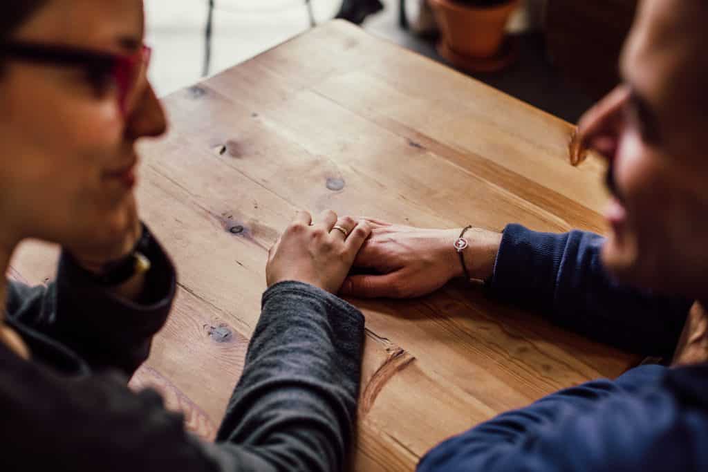 Casal conversando, com as mãos dadas sobre uma mesa de madeira.