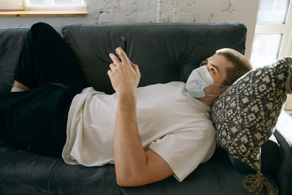 Homem deitado em sofá, usando máscara no rosto, e digitando em um celular.