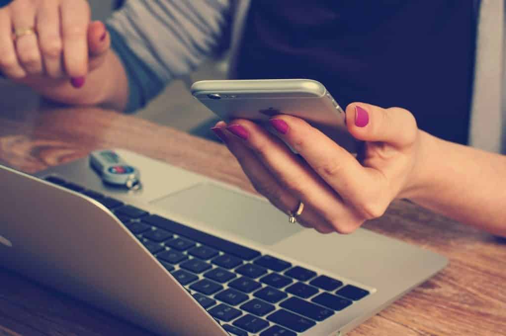 Mãos de mulher em frente a um notebook, e segurando um celular.