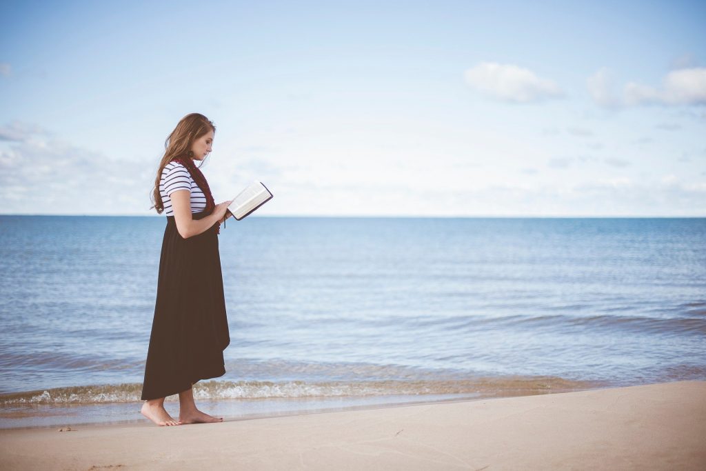 Imagem do mar e uma mulher caminhando na beira da praia com um livro aberto em suas mãos.