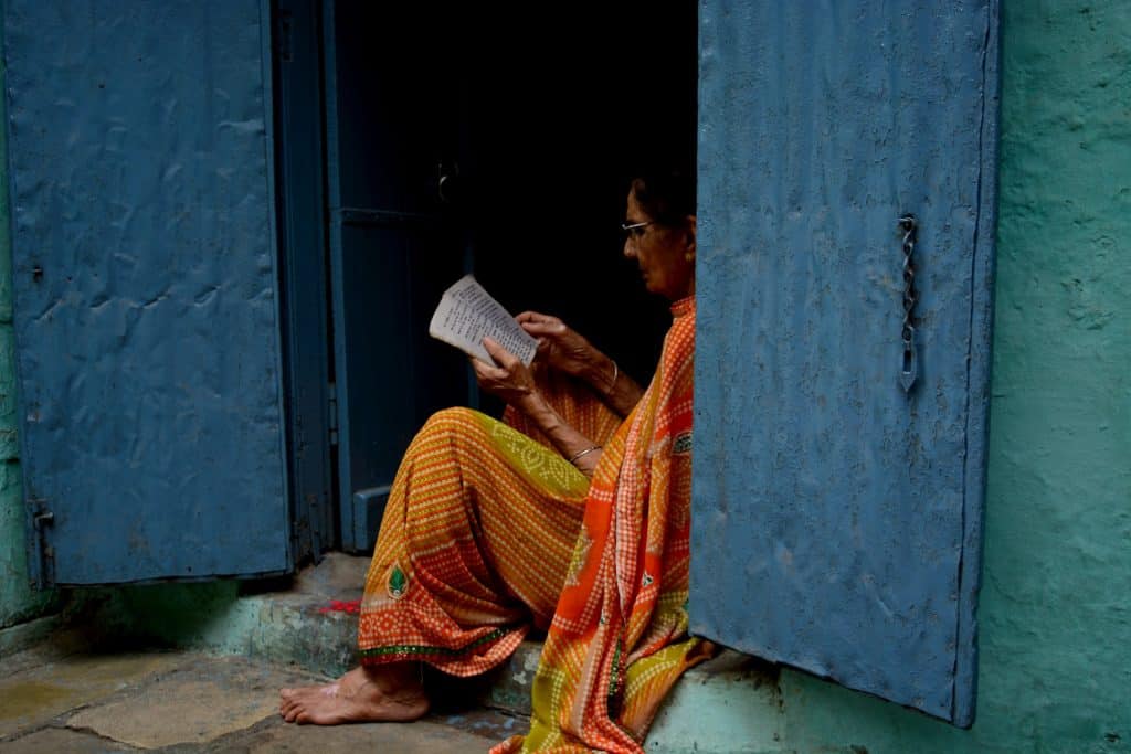 Senhora indiana sentada na soleira da porta lendo um livro.
