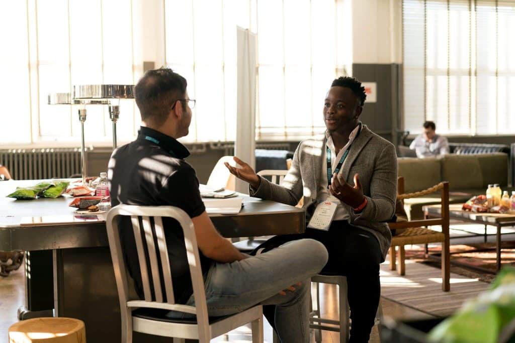 Dois homens com crachás de empresa sentados em uma mesa, conversando.