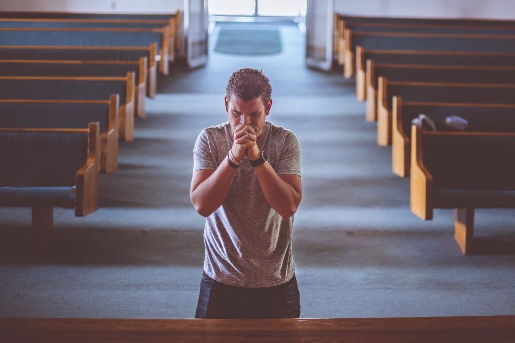 Homem dentro de um templo religioso. Ele está de joelhos orando e com os olhos fechados.