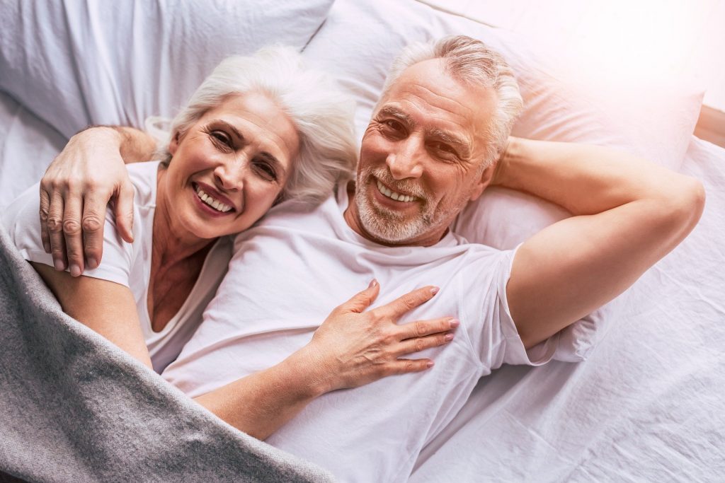 Imagem de um casal de idosos (homem e mulher) deitados na cama. Ele abraça a mulher e os dois sorriem muito, Estão felizes. Ambos usam camiseta branca e estão cobertos com uma manta cinza clara.