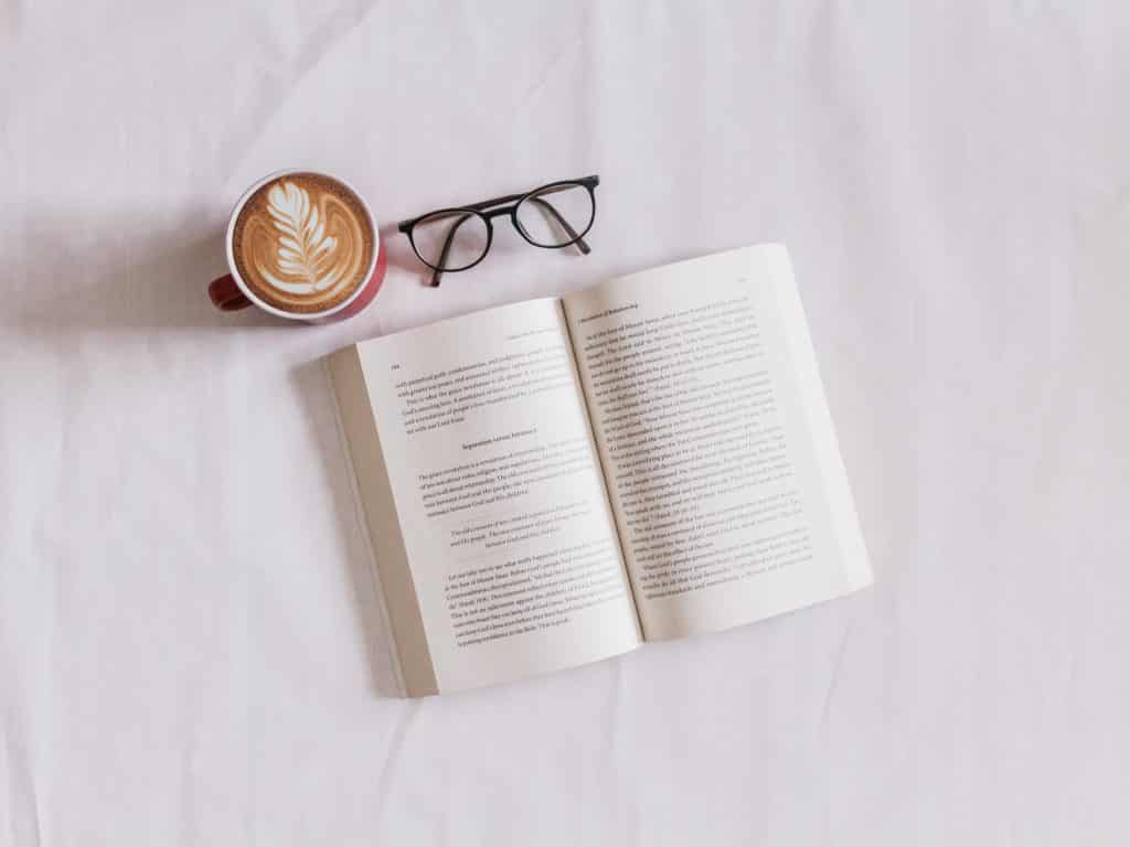 Livro aberto em uma superfície branca, ao lado está um óculos e uma caneca com café espumoso. 