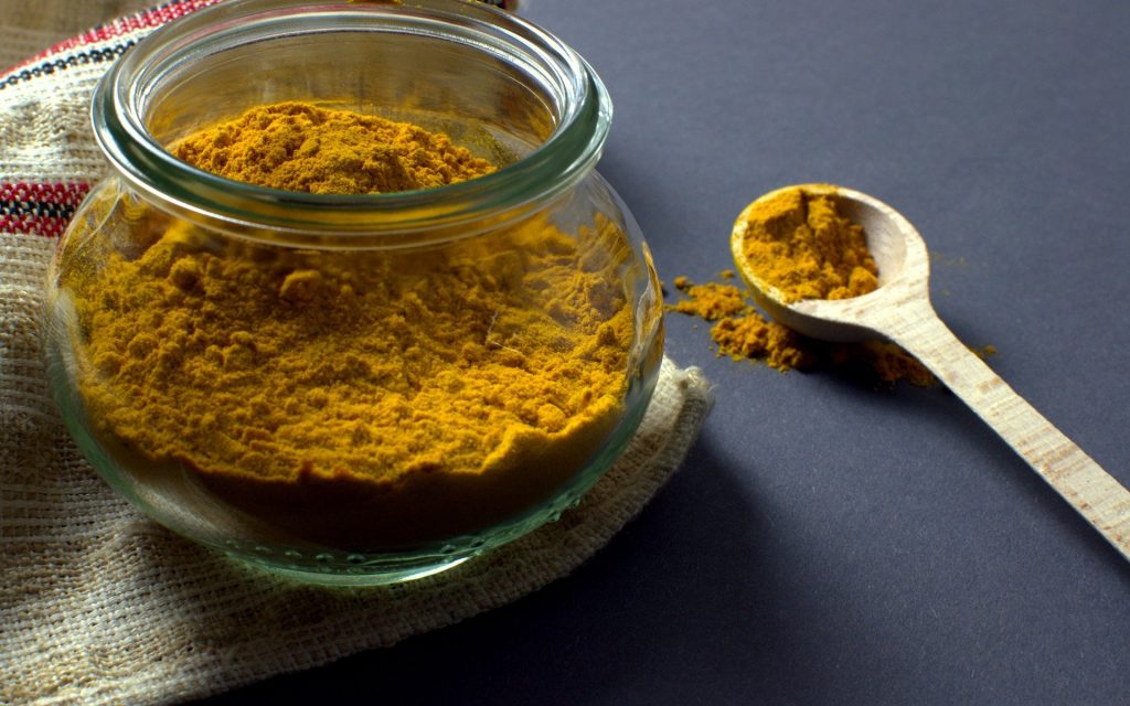 Imagem de pó de cúrcuma em um recipiente de vidro. Ao lado uma colher de madeira com um pouco do pó que será usado em uma receita natural de remédio para vitiligo.