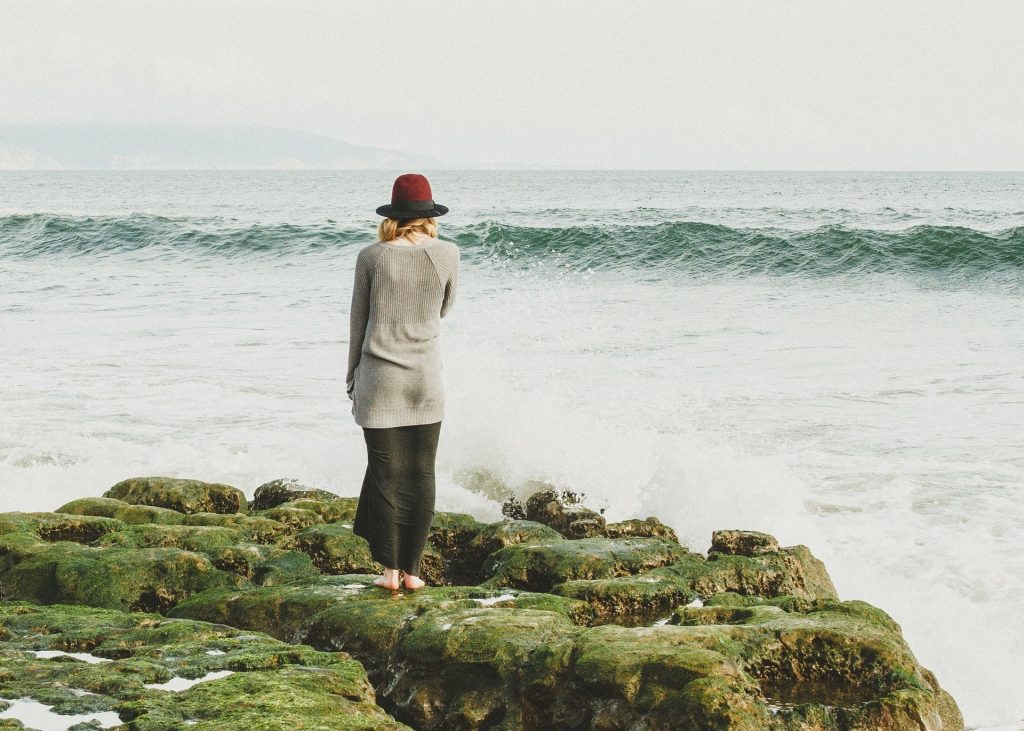 Imagem de uma mulher olhando para frente do mar em  uma tarde fria de outono. Ela veste saia longa e um cardigan cinza e um chapéu na cor vermelha.
