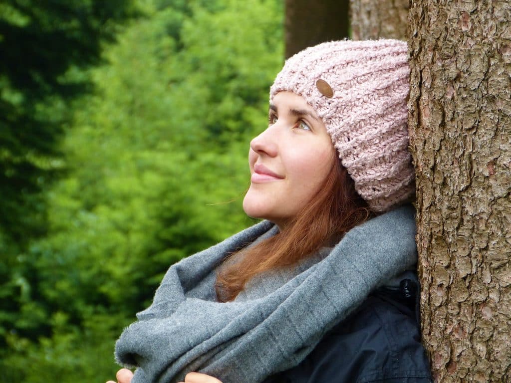 Imagem de uma jovem mulher em uma floresta. Ela está encostada em uma árvore. Está com a fisionomia alegre e relaxada. Ela usa uma trouca de lã rosa e um cachecol cinza.