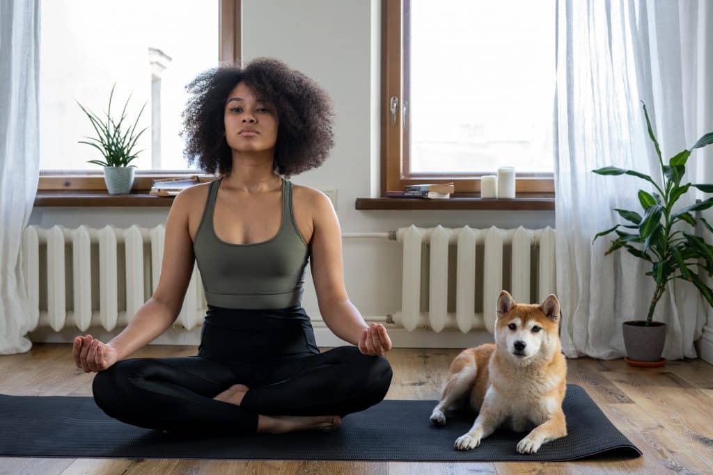 Mulher de cabelos cacheados sentada no chão da sala, ao lado de seu cachorro, meditando.