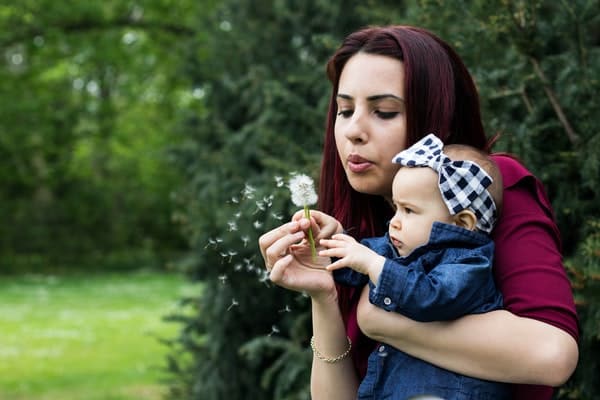 Mãe segurando filha e assoprando flor dente-de-leão