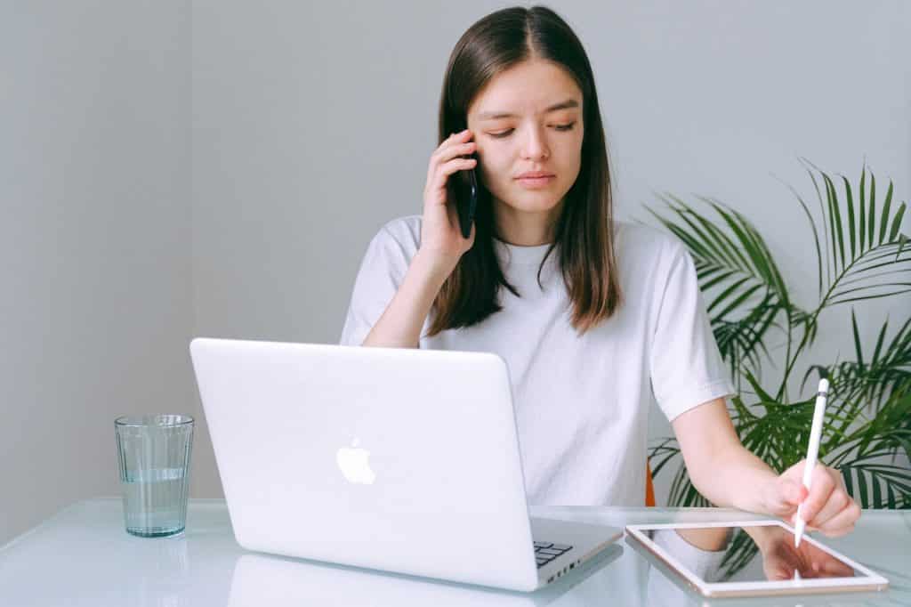 Mulher sentada em frente a um notebook e um tabet. Ela está falando em um telefone, e escrevendo no tablet com uma caneta.