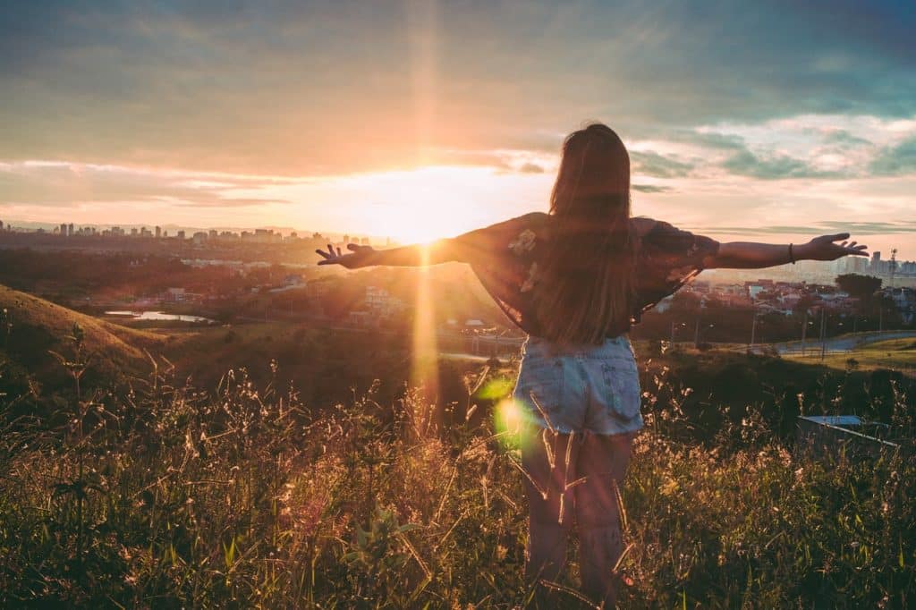 Mulher em um campo gramado, com os braços abertos para o pôr-do-sol.