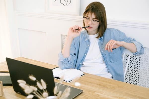 Mulher estudando sentada em cadeira com notebook e caderno na mesa e caneta na boca