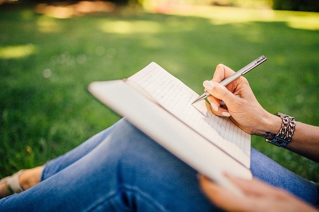 Mulher sentada em grama, com os joelhos levemente flexionados, e um caderno apoiado neles. Ela escreve no caderno com uma caneta.