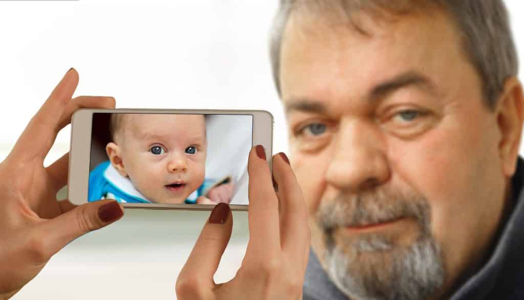 Imagem do rosto de um senhor grisalho e olhos azuis. Ele usa cavanhaque e bigode. As mãos de uma mulher segura o celular e a imagem que reflete na tela é de um bebê se referindo ao senhor quando criança.
