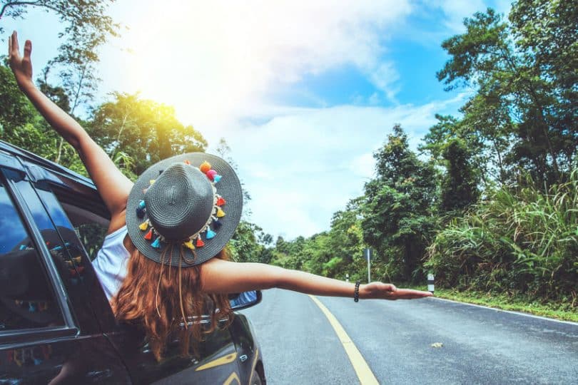 Mulher de braços abertos na janela do carro com estrada ao fundo