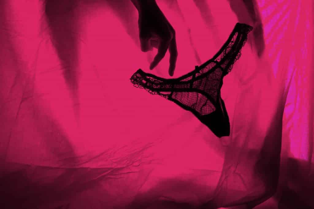 Imagem de fundo com um tecido de cetim na cor rosa escuro, Ao fundo uma mão feminina soltando uma calcinha de preta de rendinha.