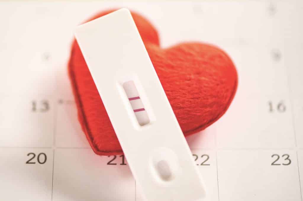 Foto de teste de gravidez positivo e junto a imagem de um calendário. Sobre ele um coração vermelho feito de feltro.