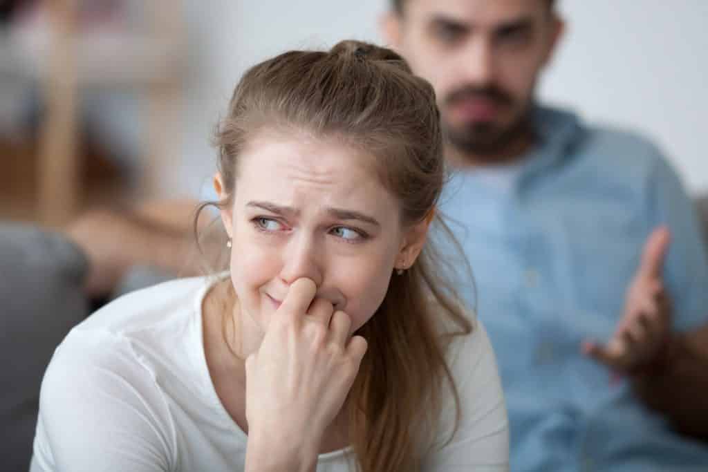 Mulher sentada no sofá, chorando, enquanto um homem gesticula atrás com expressão agressiva.