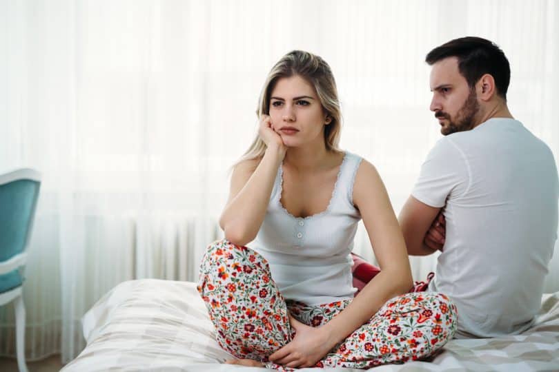 Mulher sentada na cama ao lado do marido. Ambos estão com cara de bravos.