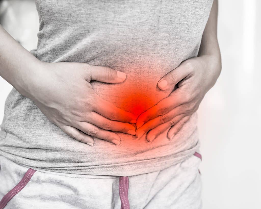 Imagem do corpo de uma mulher do busto para baixo. Ela veste uma camiseta cinza e uma calça de moletom. Aparentemente está com dores na região da barriga. Ela foi diagnosticada com útero bicorno.