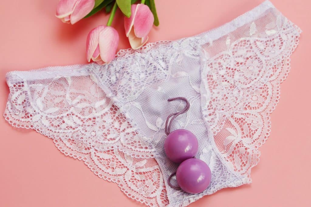 Imagem de um fundo rosa com uma calcinha branca de renda. Ao fundo botões de rosa na cor rosa e bolas chinesas complemetam a imagem.