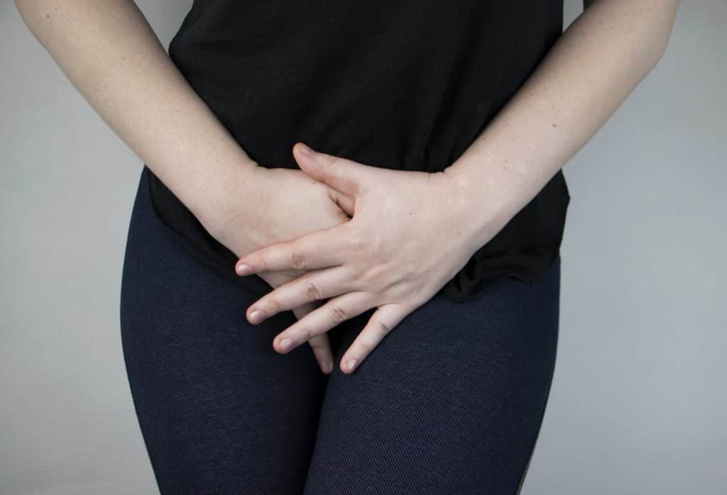 Imagem do corpo de uma mulher - do seio para baixo. Ela usa uma camiseta preta e calça jeans e está com as duas mãos sobre o seu útero. Ela está doente e o diagnóstico é mioma no útero.