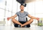 Homem sentado no chão, com as pernas cruzadas, meditando em espaço fechado, sem móveis, e com paredes de janelas.