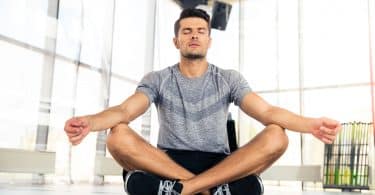 Homem sentado no chão, com as pernas cruzadas, meditando em espaço fechado, sem móveis, e com paredes de janelas.