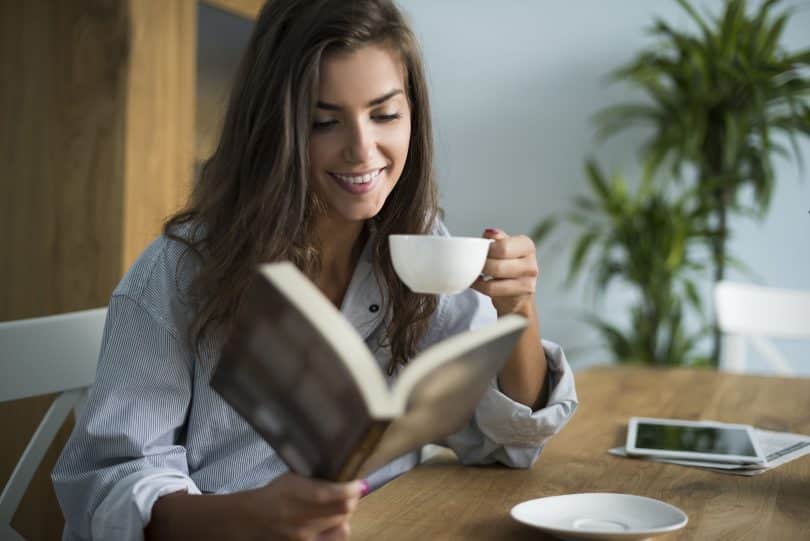 Mulher em sua mesa lendo livro e tomando café