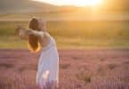 Mulher de perfil com braços abertos em campo com sol refletindo