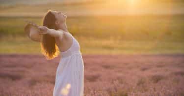 Mulher de perfil com braços abertos em campo com sol refletindo