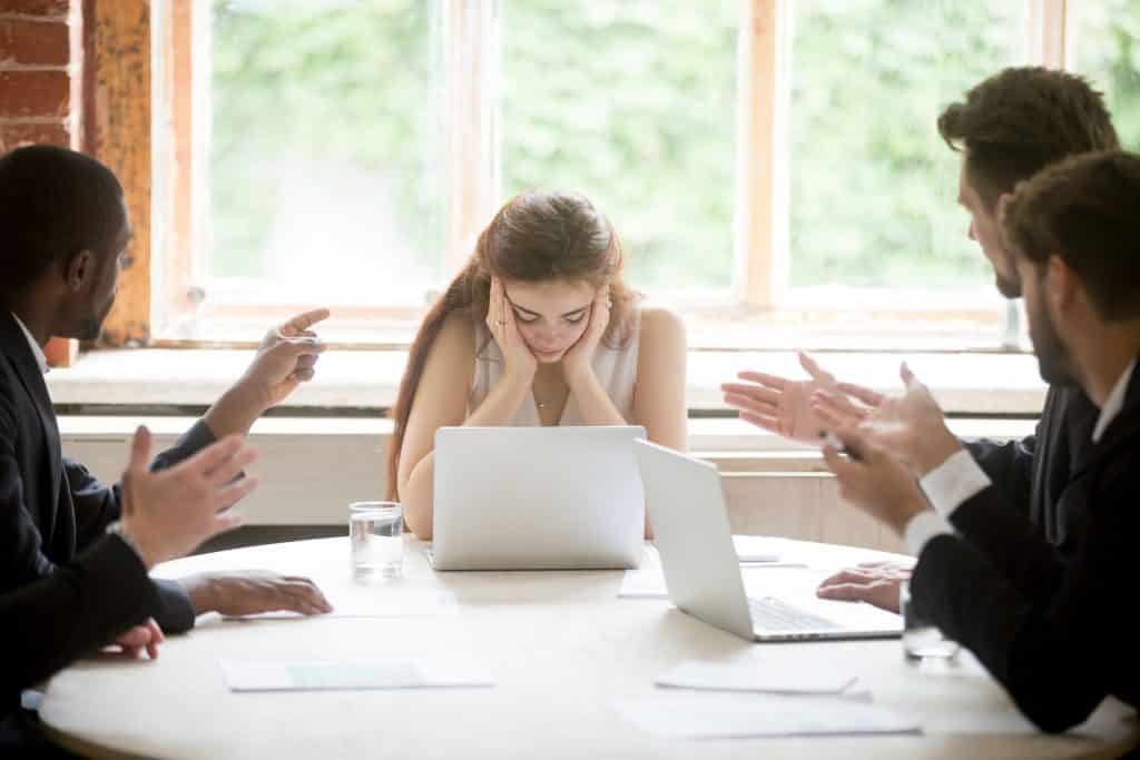 Homens discutindo com uma mulher, que segura a cabeça com as mãos, em uma mesa de reunião de esritório.