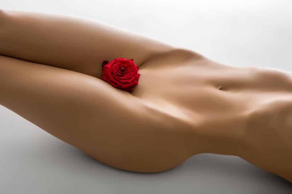 Imagem nú do orpo de uma mulher - do pescoço para baixo. Ela está com um botão de rosa vermelha entre as pernas, próximo à sua região sexual.