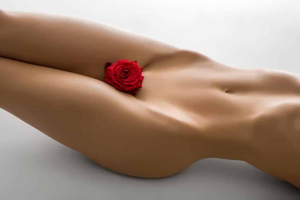 Imagem nú do orpo de uma mulher - do pescoço para baixo. Ela está com um botão de rosa vermelha entre as pernas, próximo à sua região sexual.
