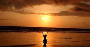 Imagem de uma mulher sentada em frente ao mar ao pôr do sol com os braços erguidos para cima