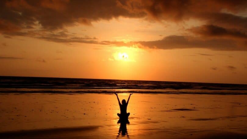 Imagem de uma mulher sentada em frente ao mar ao pôr do sol com os braços erguidos para cima