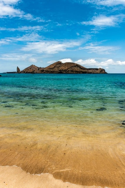 Praia com águas claras e pedras ao fundo com céu azul com nuvens