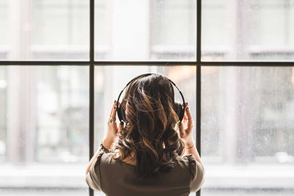 Mulher vista de costas olhando pela janela enquanto escuta música em seu fone de ouvido.