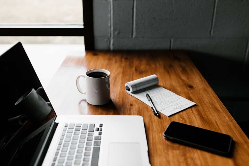 Mesa com notebook aberto e ao lado uma xícara de café junto com caderno de anotação