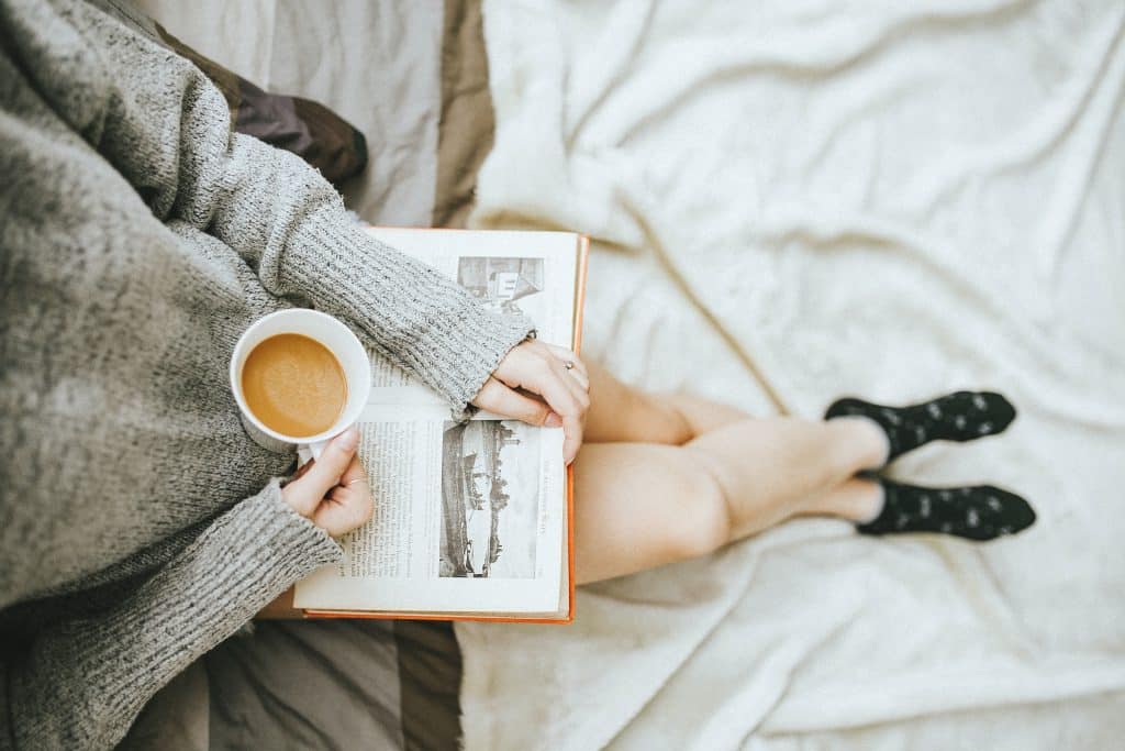 Mulher sentada na cama com livro aberto apoiado em suas pernas e segurando xícara de chá